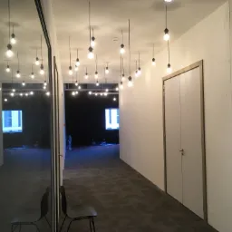 Wnętrze biurowe z zamontowanym oświetleniem wiszącym, widok na korytarz z szarym dywanem, białe ściany i drzwi, odbicie w szklanej powierzchni z krzesłami.