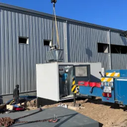 Montaż transformatora w Jeleniej Górze za pomocą dźwigu, pracownik wewnątrz stacji transformatorowej podłącza kable, widoczny budynek przemysłowy w tle.