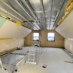 Poddasze w trakcie adaptacji: widoczne stelaże sufitu, izolacja wełną mineralną, częściowo zamontowane płyty karton-gips, szpachlowane ściany, drabina i narzędzia.