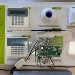 Centrala alarmowa Perfecta z manipulatorem, czujnikiem ruchu, sygnalizatorem optycznym i anteną GSM, prezentowana na planszy z opisami technicznymi.