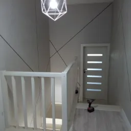 Klatka schodowa po odnowieniu z białą balustradą, drzwi z poziomymi przeszkleniami i modernistyczną lampą wiszącą, na podłodze leży wkrętarka.