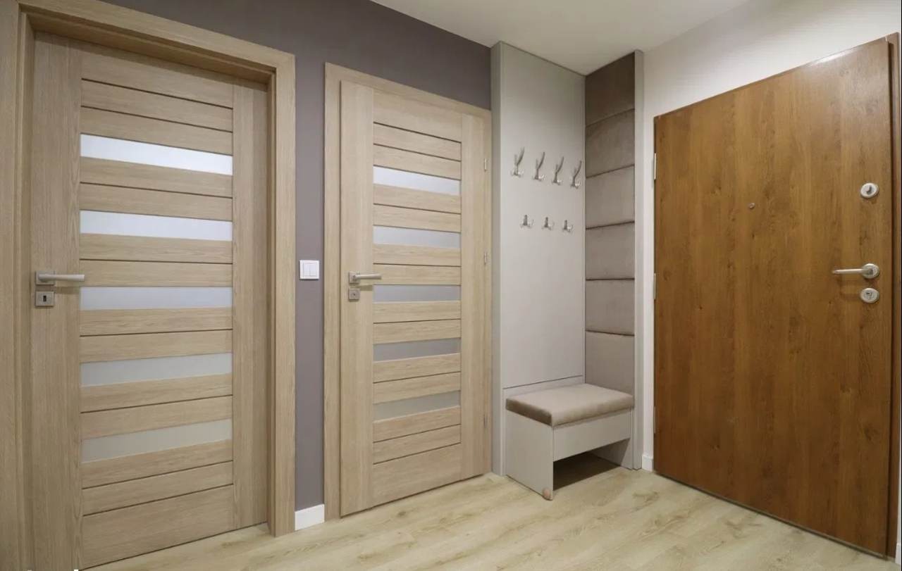 Jasne, drewniane drzwi wewnętrzne z poziomymi przeszkleniami, obok brązowe drzwi wejściowe, w tle wieszak z siedziskiem w przedpokoju.