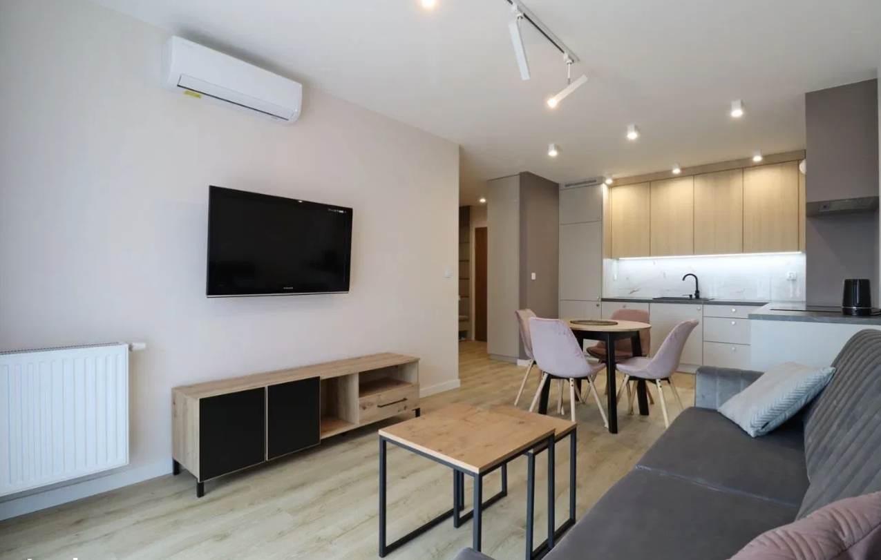 Jasne, nowocześnie urządzone wnętrze mieszkania z aneksem kuchennym, widok na salon z szarą sofą, stolikiem kawowym i szafką RTV pod wiszącym telewizorem, w tle okrągły stół z krzesłami i zabudowa...