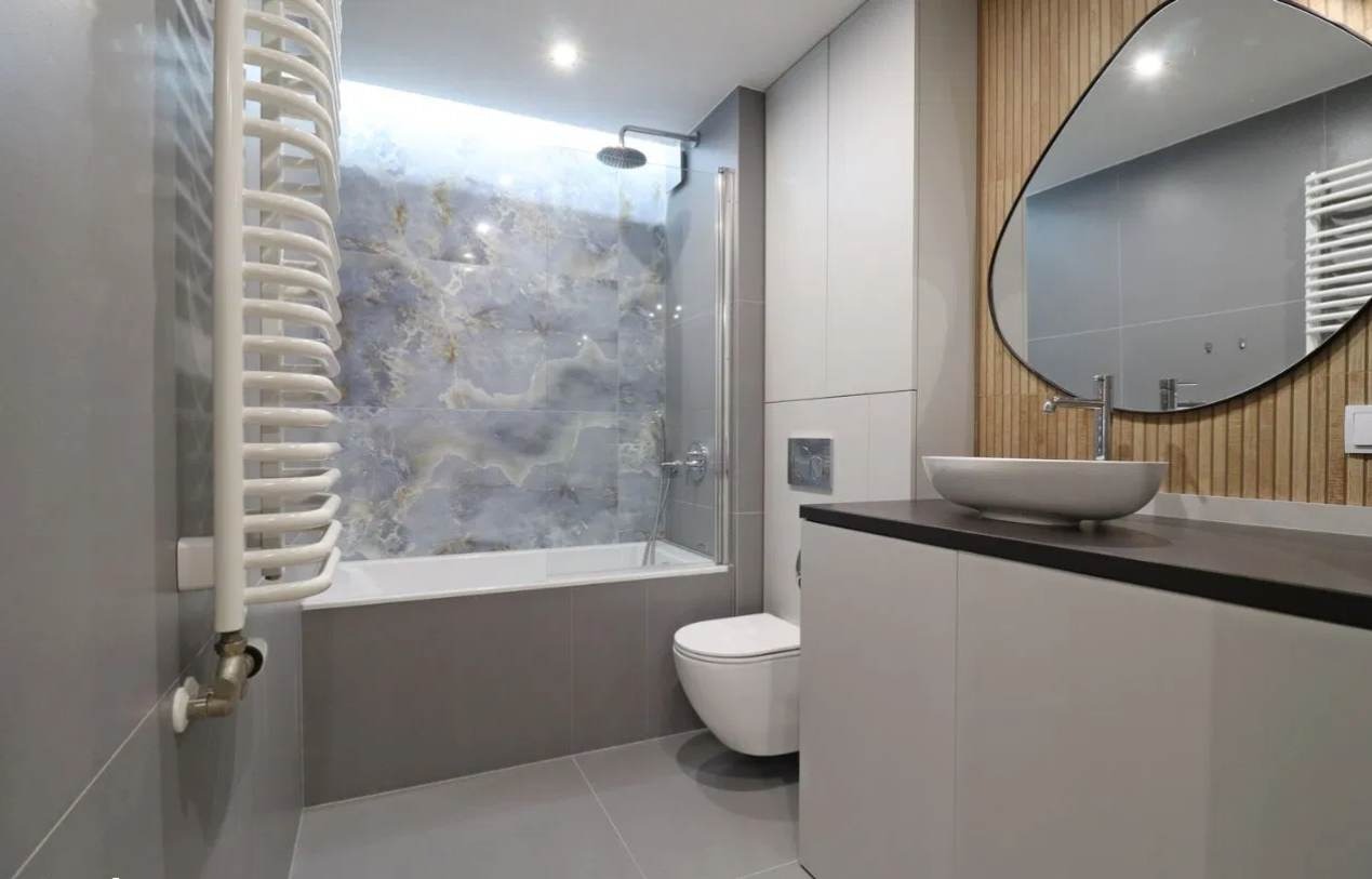 Nowoczesna łazienka w odcieniach szarości z akcentami drewna, owalne lustro nad umywalką nablatową, marmurowe płytki w strefie kąpieli z parawanem nawannowym, grzejnik drabinkowy na ścianie.