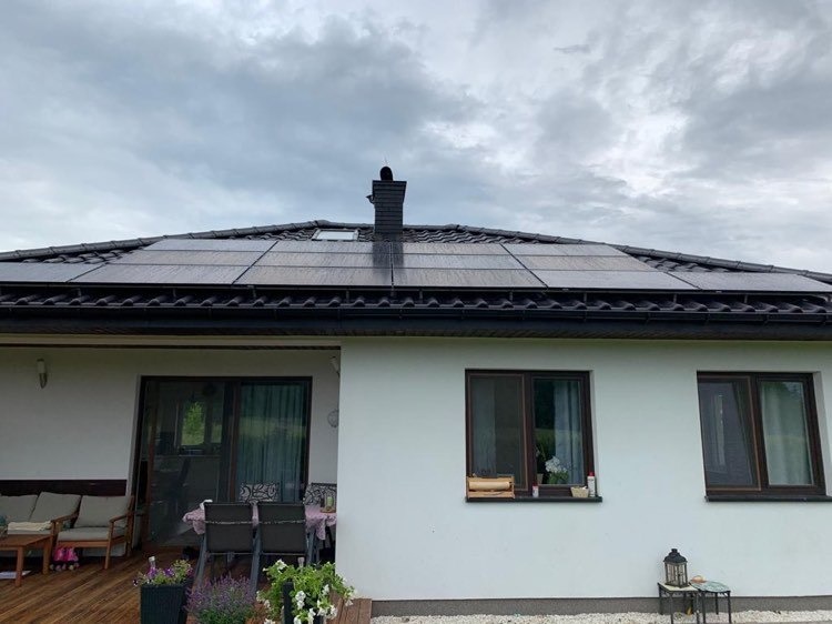 Czarny dach skośny z panelami fotowoltaicznymi, widok z poziomu tarasu z meblami ogrodowymi i kwiatami, pochmurne niebo.