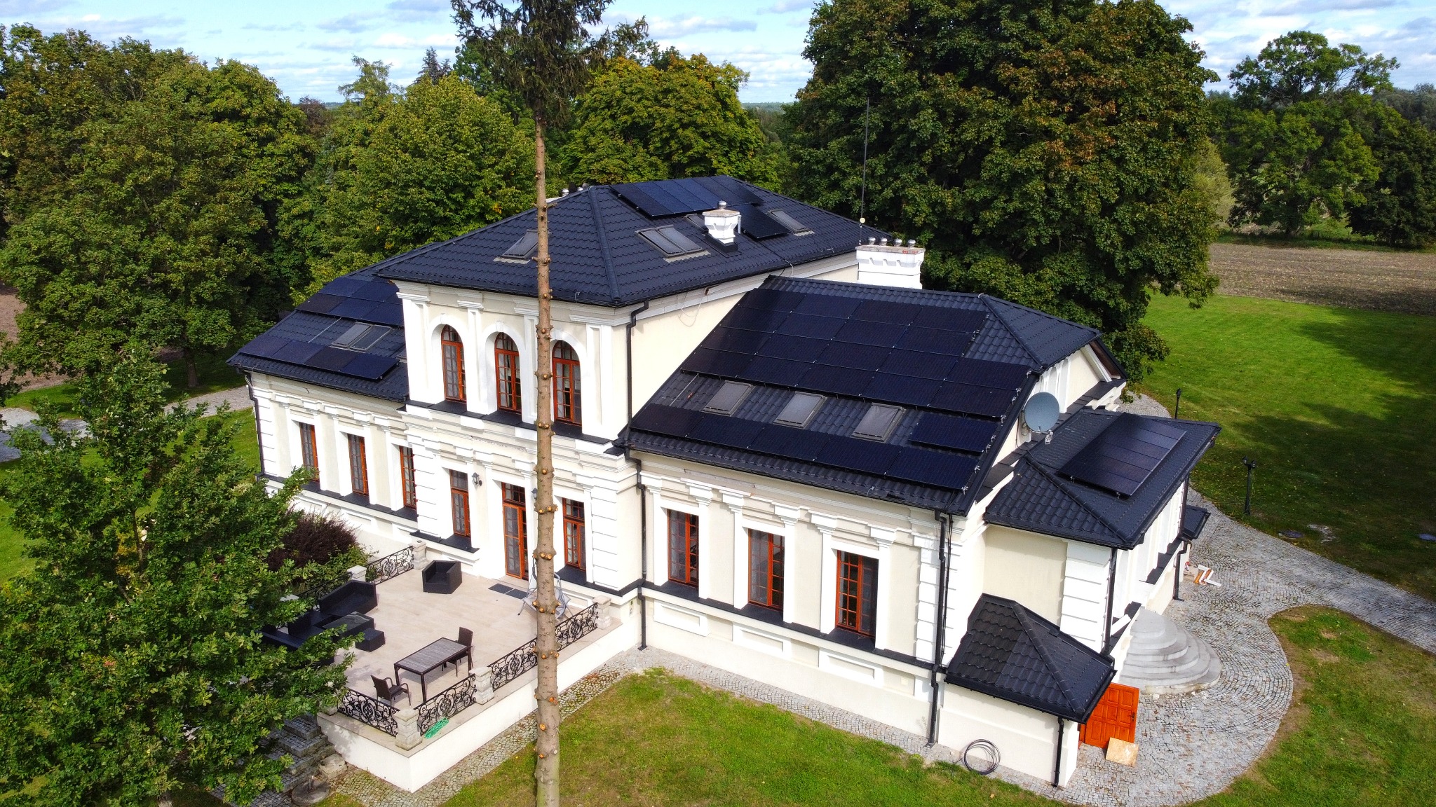 Elegancki dom z kremową elewacją i czarną dachówką, na której zamontowane są panele fotowoltaiczne i okna dachowe, otoczony zielonym trawnikiem i drzewami, z widocznym tarasem z meblami.
