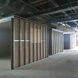 Montaż ścian działowych z płyty karton gips na metalowym stelażu w surowym stanie, widoczne instalacje pod sufitem w przestrzeni biurowej.