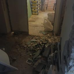 JustHome Piotr Baczewski Usługi remontowo-budowlane - Wnętrze w trakcie demontażu: gruz, fragmenty płytek, worki z materiałami budowlanymi i pracownik w tle.