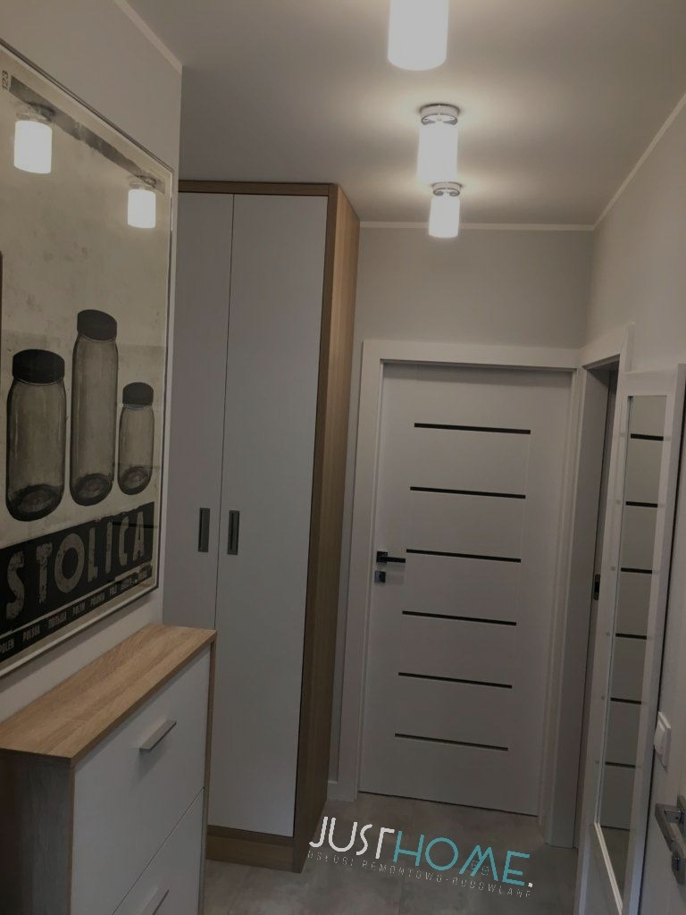 Nowoczesny przedpokój z białą szafą w zabudowie, komodą z drewnianym blatem, plakatem w ramie i białymi drzwiami z poziomymi frezami, oświetlony trzema cylindrycznymi lampami sufitowymi.