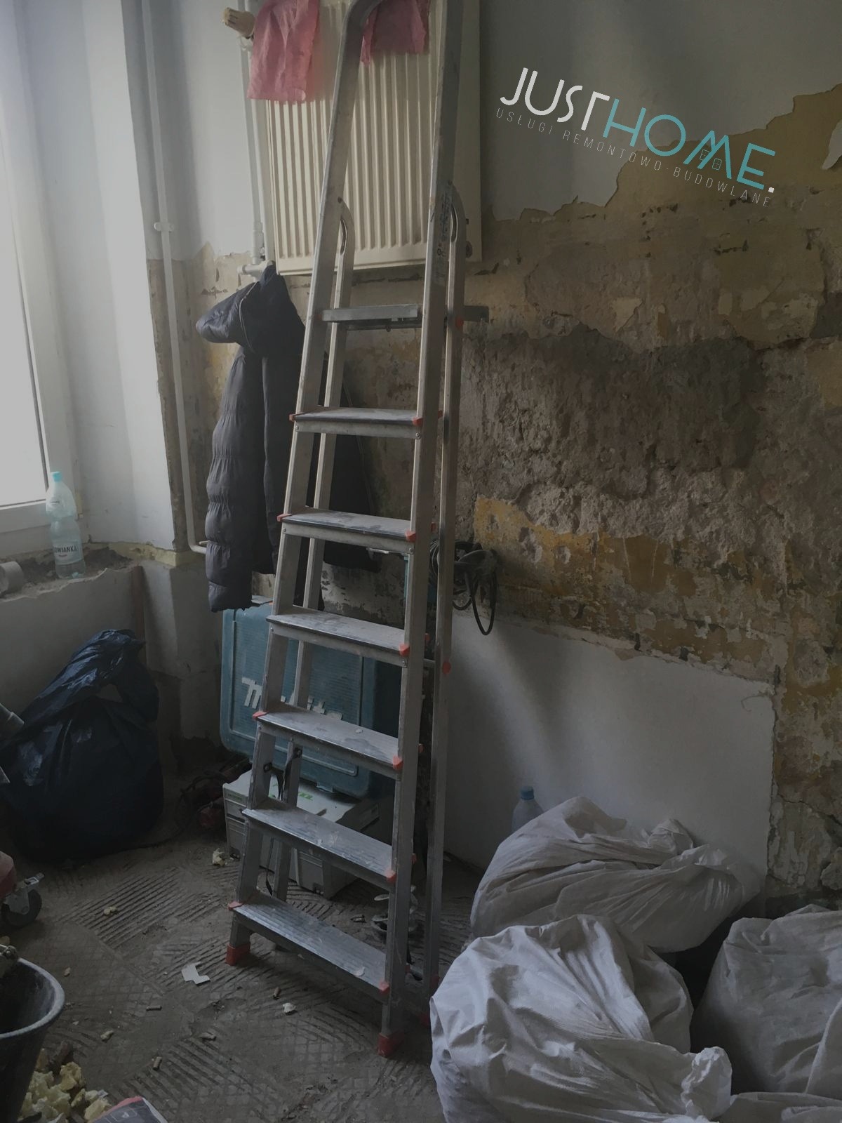 Pokój w trakcie prac przygotowawczych do malowania: ściany częściowo pokryte nową warstwą, drabina aluminiowa, worki z materiałem, narzędzia i logo firmy JustHome.