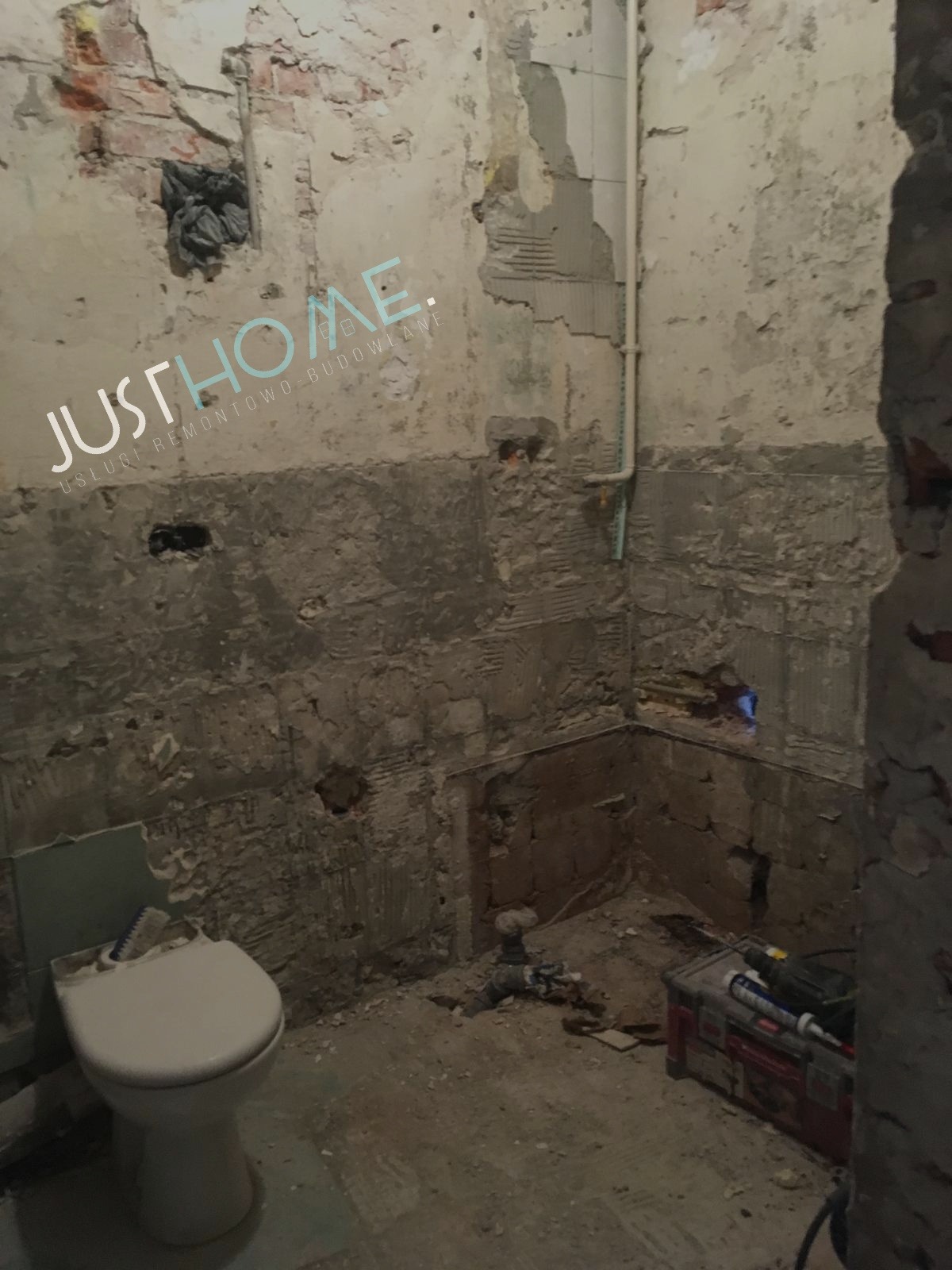 Łazienka w trakcie remontu: zdemontowane płytki, odsłonięta cegła, widoczny fragment toalety i skrzynka z narzędziami na podłodze.