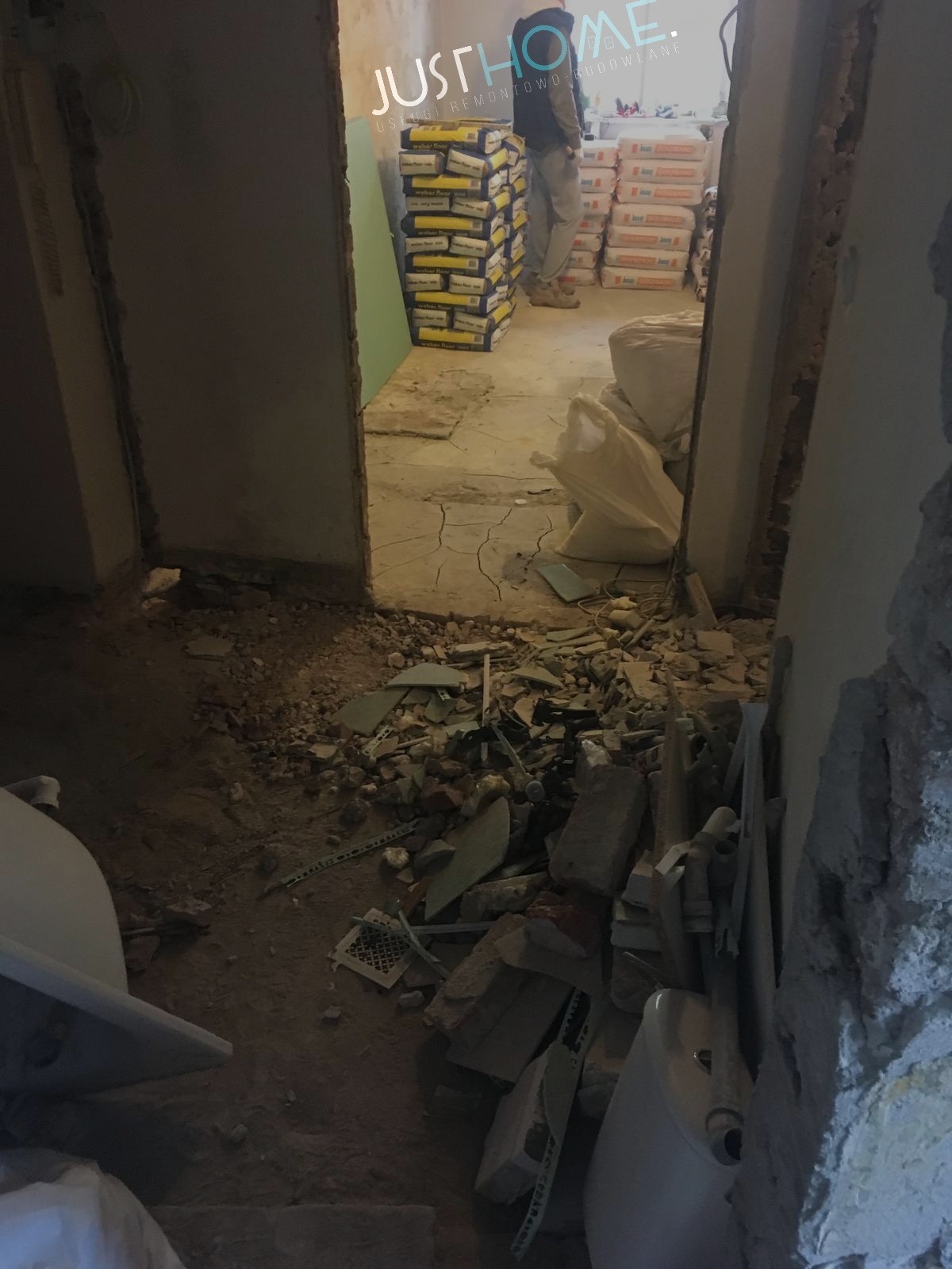 Wnętrze w trakcie demontażu: gruz, fragmenty płytek, worki z materiałami budowlanymi i pracownik w tle.