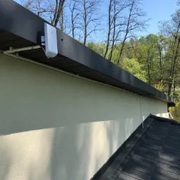 Biały punkt dostępowy WiFi zamontowany pod okapem domu z widocznymi przewodami i rurkami instalacyjnymi, tło stanowi jezioro i drzewa.