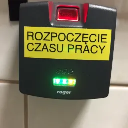 Elektroniczny czytnik zbliżeniowy firmy Roger z napisem 'Rozpoczęcie Czasu Pracy', podświetlony diodami LED na czerwono i zielono, zamontowany na jasnej ścianie.