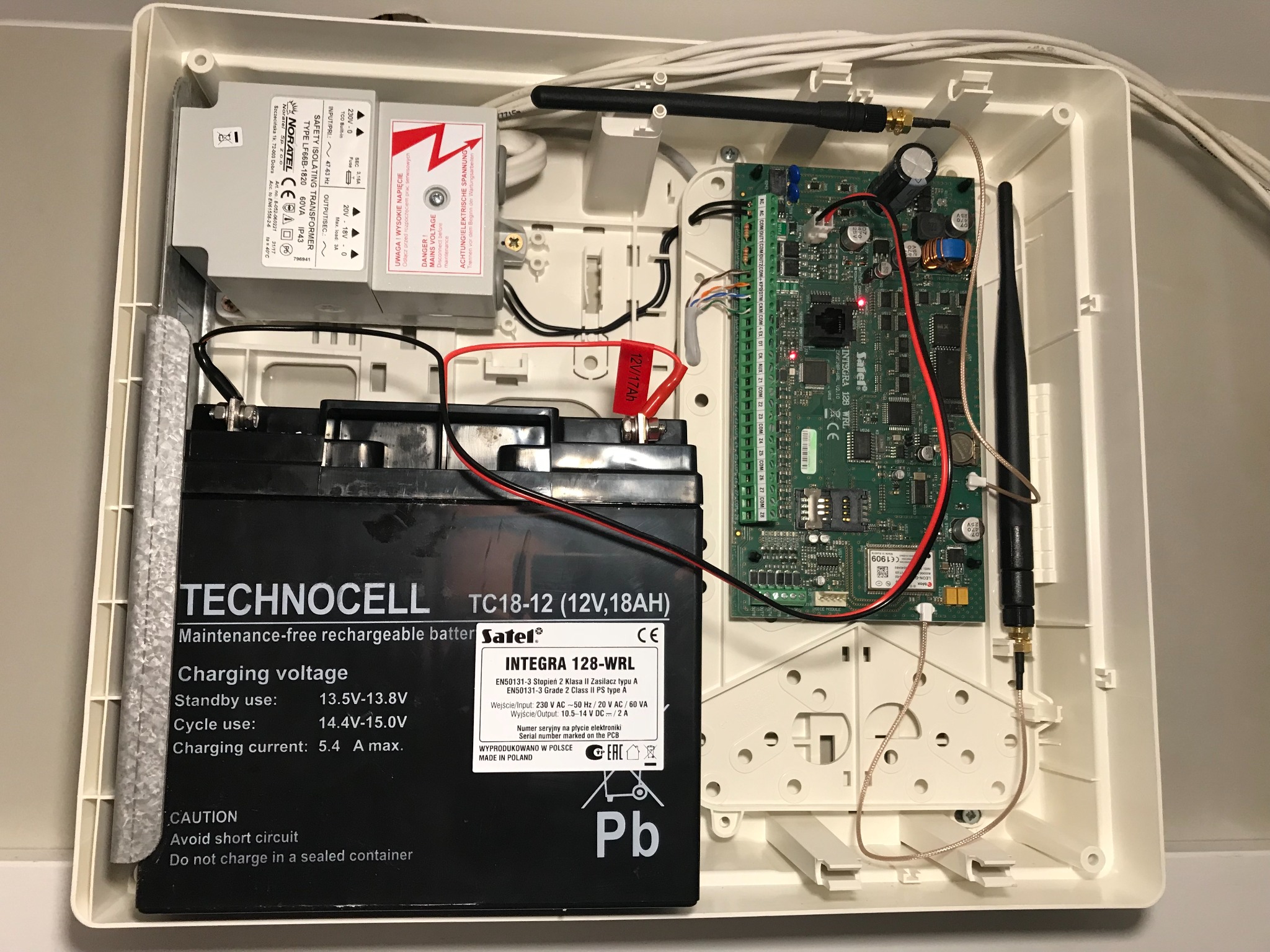 Otwarta obudowa centrali alarmowej z widoczną płytą główną, akumulatorem i transformatorem, kable połączeniowe i antena.