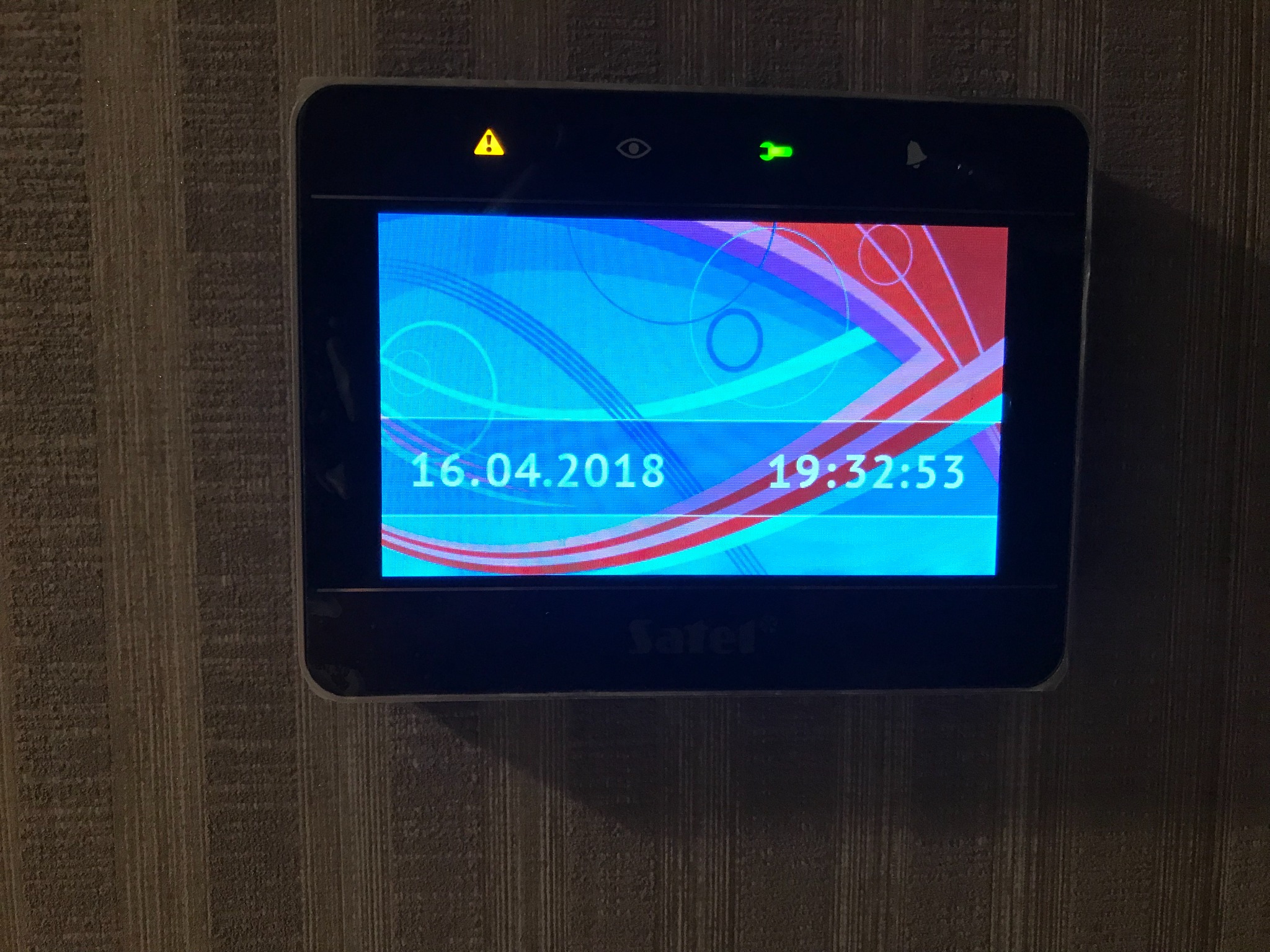 Ekran centrali alarmowej marki Satel z wyświetlaną datą i godziną, zamontowany na ścianie o fakturze pionowych pasków.
