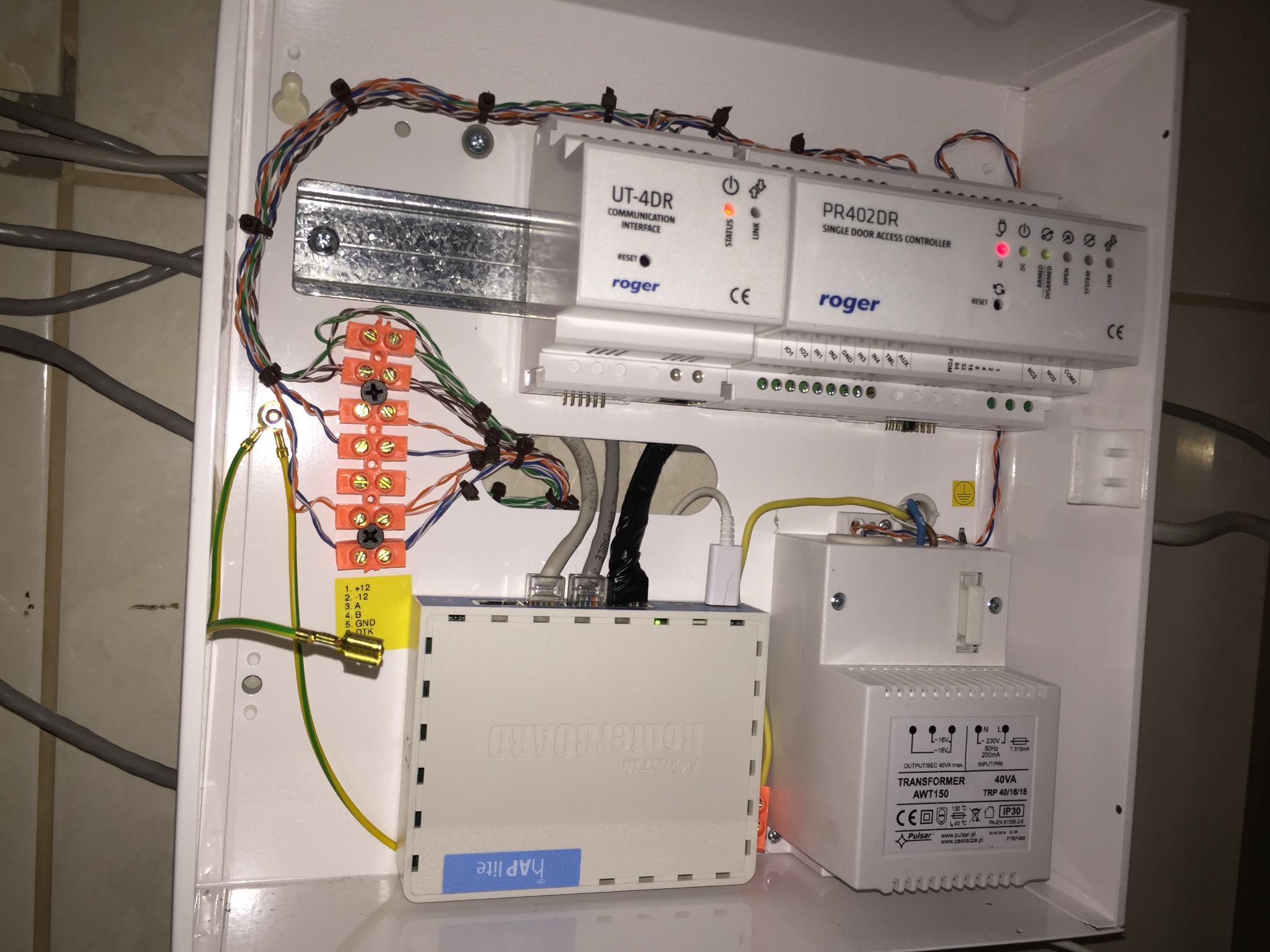 Wnętrze białej skrzynki z zamontowanymi urządzeniami: kontroler dostępu Roger PR402DR, interfejs komunikacyjny Roger UT-4DR, router MikroTik hAP lite i transformator Pulsar AWT150, połączone kablami.