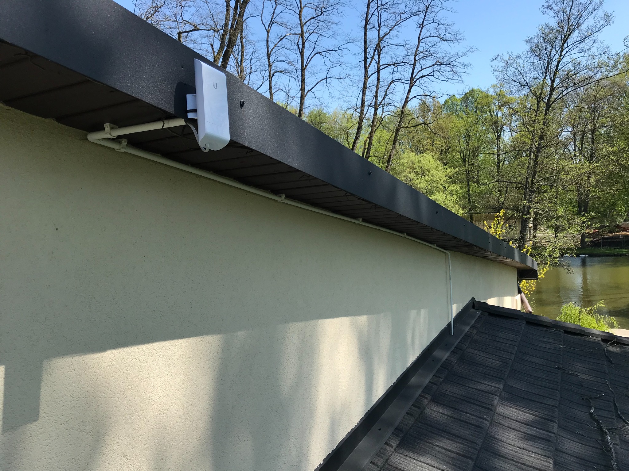 Biały punkt dostępowy WiFi zamontowany pod okapem domu z widocznymi przewodami i rurkami instalacyjnymi, tło stanowi jezioro i drzewa.