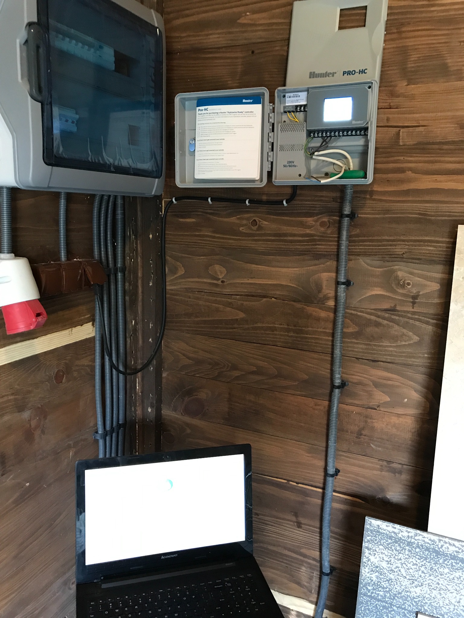 Konfiguracja sterownika nawadniania Hunter Pro-HC za pomocą laptopa na tle drewnianej ściany, widoczne przewody w peszlach i skrzynka elektryczna.