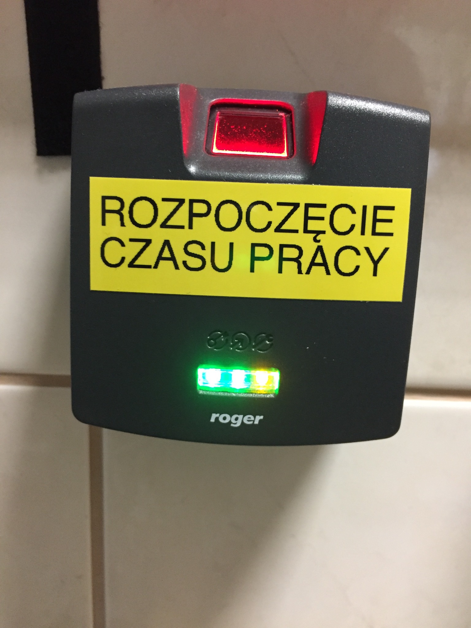 Elektroniczny czytnik zbliżeniowy firmy Roger z napisem 'Rozpoczęcie Czasu Pracy', podświetlony diodami LED na czerwono i zielono, zamontowany na jasnej ścianie.