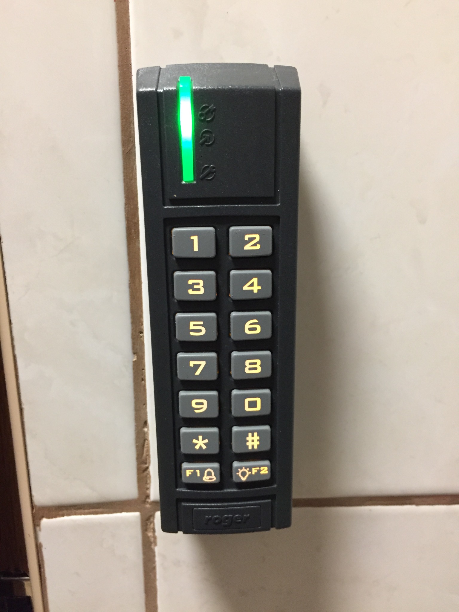 Czarny panel numeryczny Roger z zieloną diodą LED, zamontowany na kafelkach ściennych, widoczne przyciski z cyframi i symbolami.