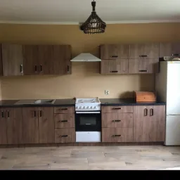 Wykończona kuchnia z brązowymi szafkami o drewnianej fakturze, czarnym blatem i białą lodówką, oświetlona lampą z wikliny.