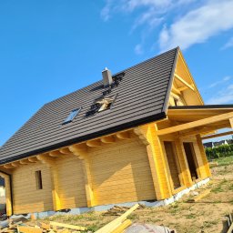 WILK-BUD SPÓŁKA Z OGRANICZONĄ ODPOWIEDZIALNOŚCIĄ - Surowy, drewniany dom szkieletowy z ciemnoszarą dachówką, widoczne okno dachowe i komin, na dachu zamontowane stopnie kominiarskie, na ziemi leżą deski, w tle błękitne niebo i samochód dostawczy.