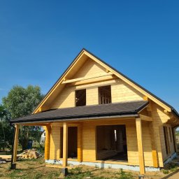 WILK-BUD SPÓŁKA Z OGRANICZONĄ ODPOWIEDZIALNOŚCIĄ - Surowy, drewniany dom szkieletowy w trakcie budowy, z widoczną konstrukcją dachu pokrytą czarną papą, otoczony zielenią i błękitnym niebem.
