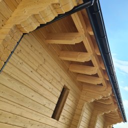 WILK-BUD SPÓŁKA Z OGRANICZONĄ ODPOWIEDZIALNOŚCIĄ - Surowa drewniana konstrukcja domu szkieletowego z widoczną więźbą dachową i czarną rynną na tle błękitnego nieba.