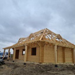 WILK-BUD SPÓŁKA Z OGRANICZONĄ ODPOWIEDZIALNOŚCIĄ - Surowy stan budowy domu z bali, widoczna konstrukcja drewniana dachu i ściany, na tle pochmurnego nieba, obok zaparkowany biały bus z logo firmy.