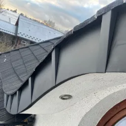 Detale czarnego, metalowego wykończenia dachu z widocznym oświetleniem zewnętrznym wbudowanym w elewację budynku. W tle budynki pokryte śniegiem.