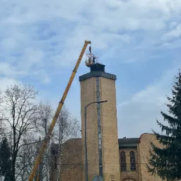 Prace na szczycie ceglanej wieży kościelnej wykonywane z podnośnika koszowego, widoczne narzędzia i robotnicy na wysokości.