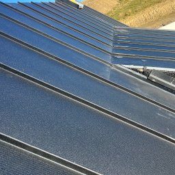 WILK-BUD SPÓŁKA Z OGRANICZONĄ ODPOWIEDZIALNOŚCIĄ - Nowe, czarne, metalowe pokrycie dachowe z widoczną konstrukcją komina w trakcie montażu na tle wzgórza i błękitnego nieba.