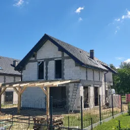 Surowy stan domu z białego bloczka z czarną dachówką, widoczna drewniana konstrukcja zadaszenia tarasu, drabina oparta o ścianę i częściowo ogrodzony teren budowy w słoneczny dzień.