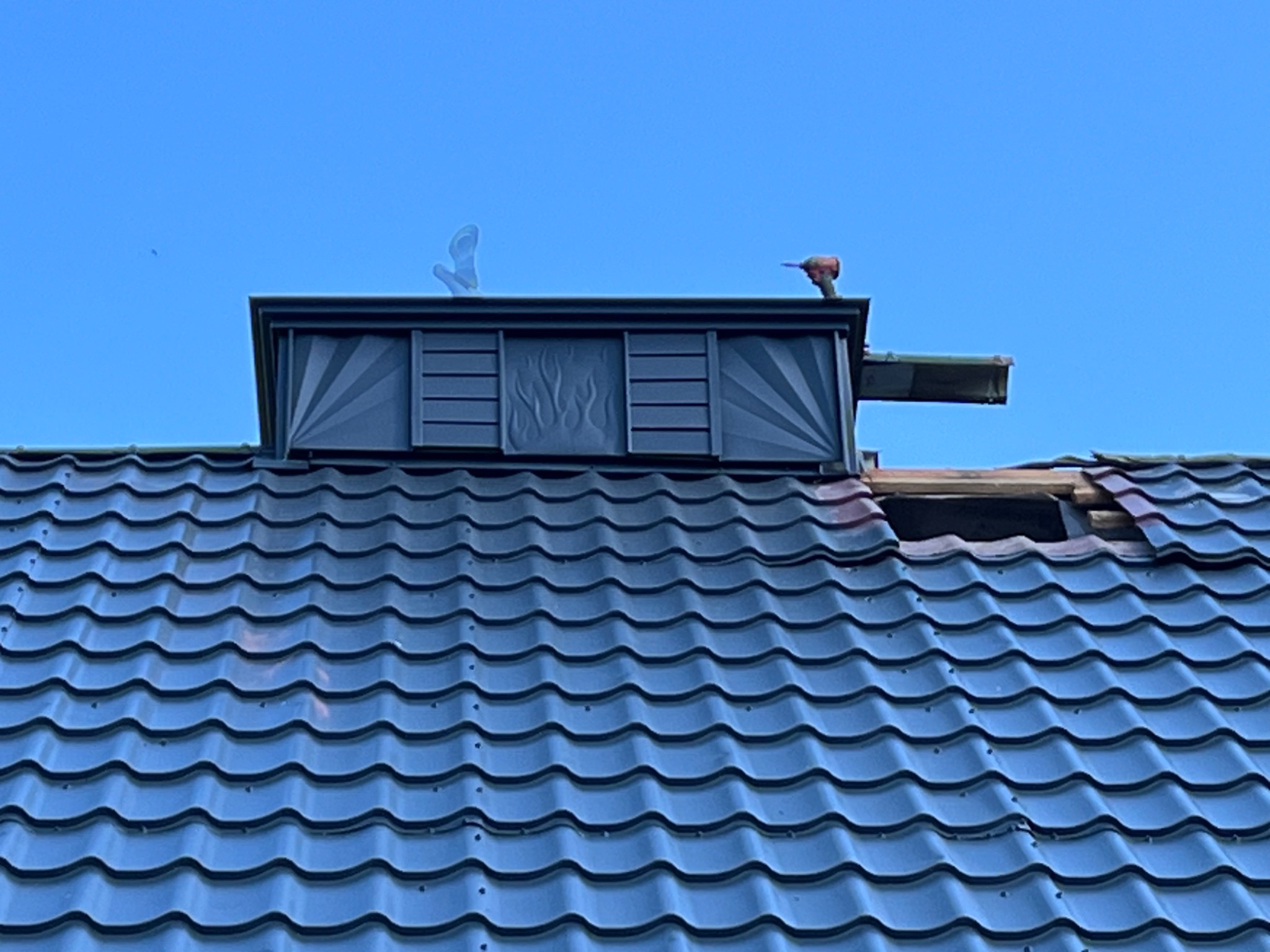 Niebieski dach z blachodachówki w Odrowążu, z kominem ozdobionym motywem płomieni i narzędziem elektrycznym na krawędzi, fragment dachu uszkodzony.