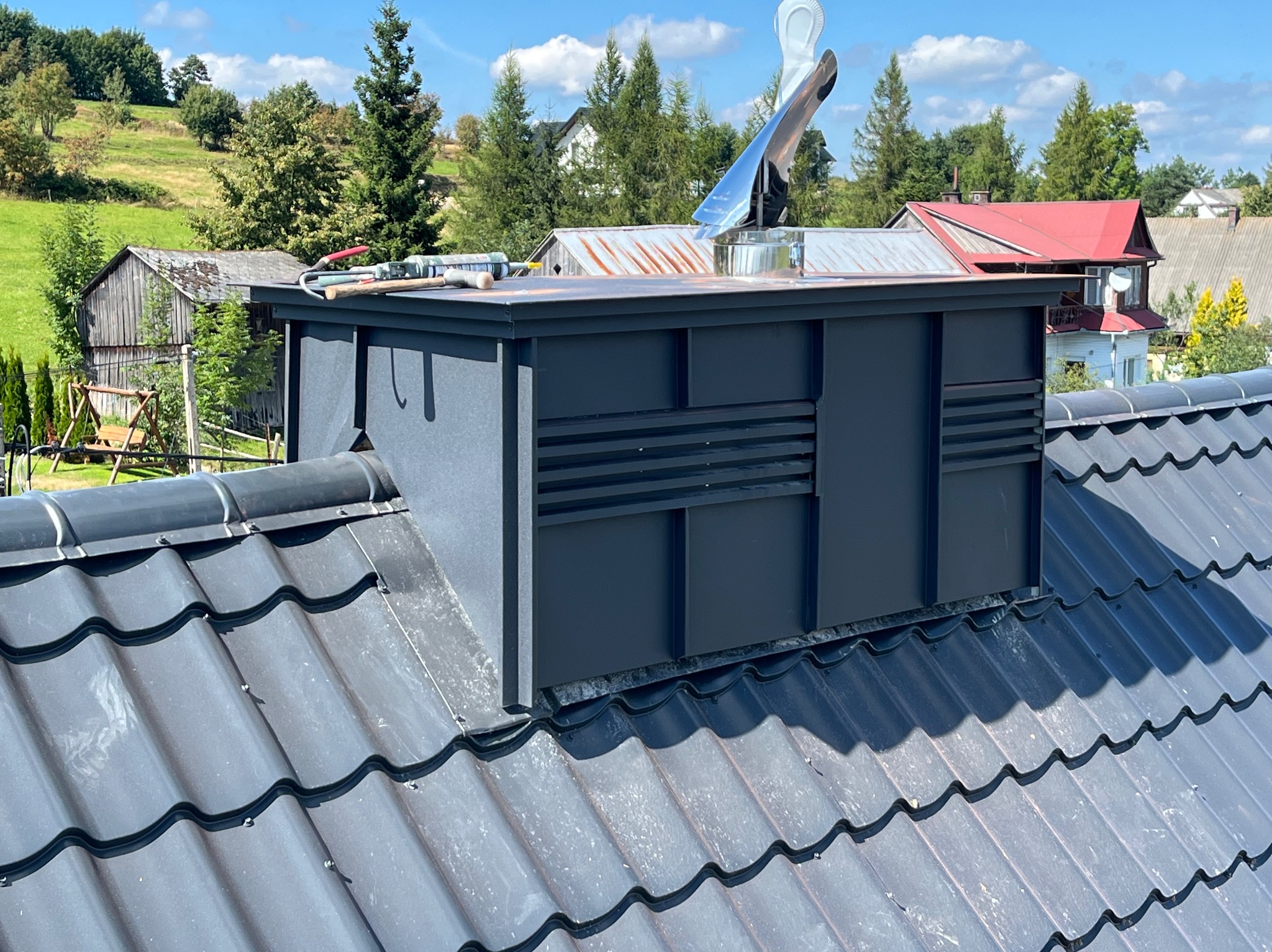 Nowoczesny, antracytowy komin wentylacyjny z metalowym wiatrołapem na dachu z blachodachówki w odcieniach szarości, z widocznymi narzędziami dekarskimi i otoczeniem wiejskim w tle.