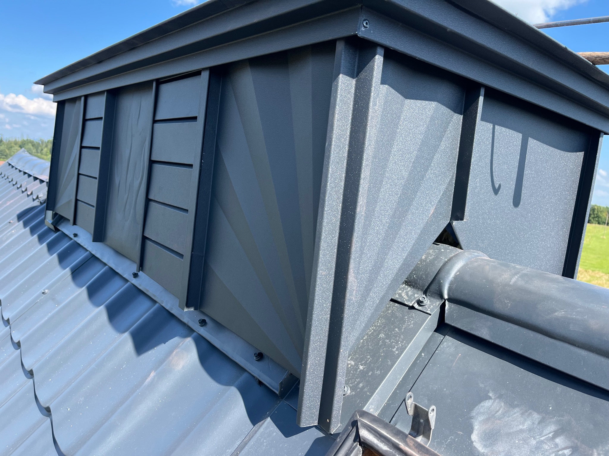 Szary komin wentylacyjny z metalu na dachu pokrytym szarą blachodachówką, widok z dołu pod kątem, na tle błękitnego nieba.