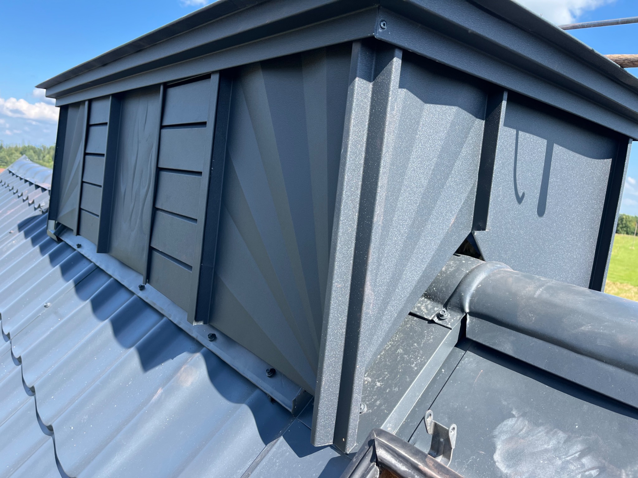 Szary komin wentylacyjny z poziomymi żaluzjami na metalowym dachu z dachówki modułowej, widoczne mocowania śrubowe i elementy obróbki blacharskiej.