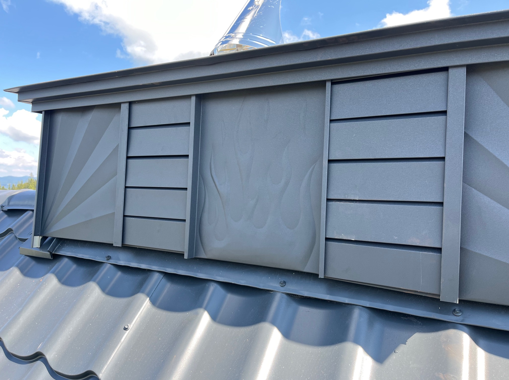 Szary odpowietrznik dachowy z dekoracyjnymi panelami: promieniste linie z lewej, płomienie pośrodku, poziome listwy z prawej, na tle błękitnego nieba.