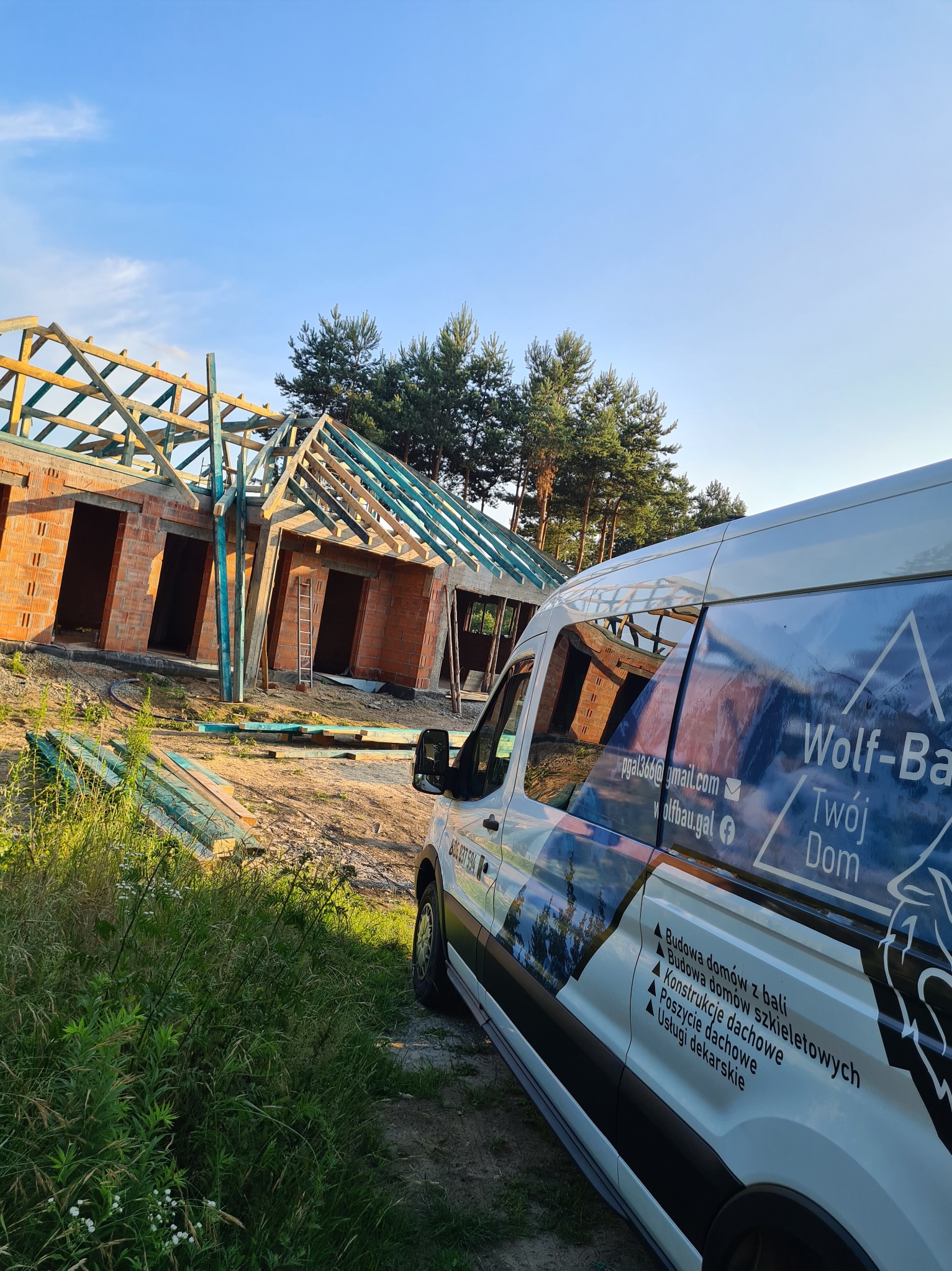 Budowa domu w trakcie z widoczną więźbą dachową i murem z cegły, na pierwszym planie samochód firmowy z logo i informacjami kontaktowymi.