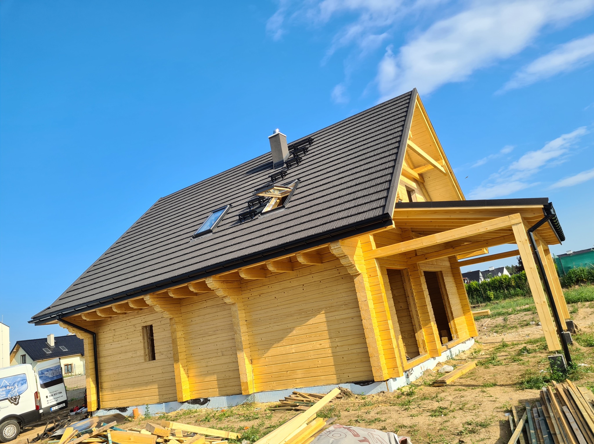 Surowy, drewniany dom szkieletowy z ciemnoszarą dachówką, widoczne okno dachowe i komin, na dachu zamontowane stopnie kominiarskie, na ziemi leżą deski, w tle błękitne niebo i samochód dostawczy.