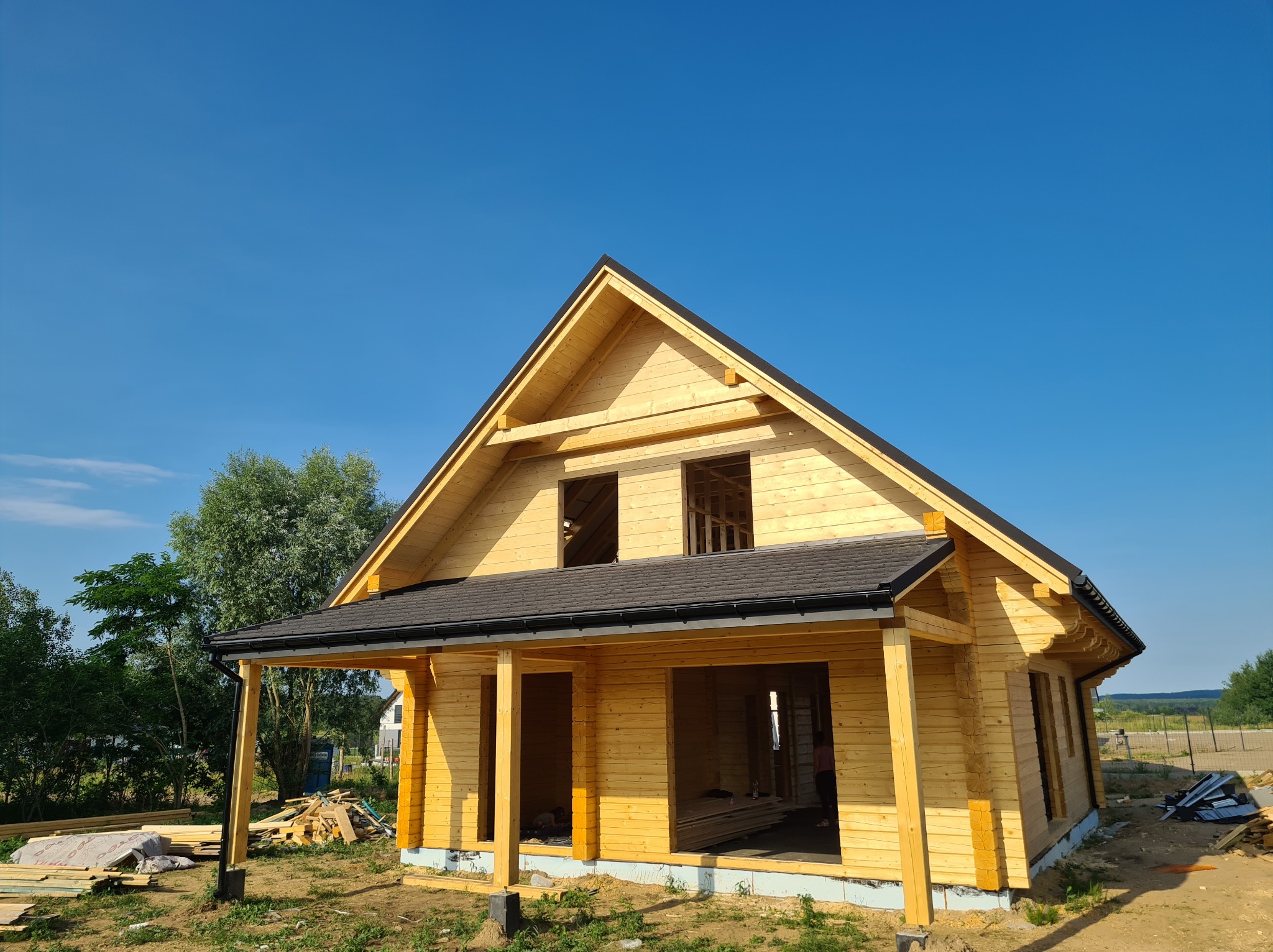 Surowy, drewniany dom szkieletowy w trakcie budowy, z widoczną konstrukcją dachu pokrytą czarną papą, otoczony zielenią i błękitnym niebem.