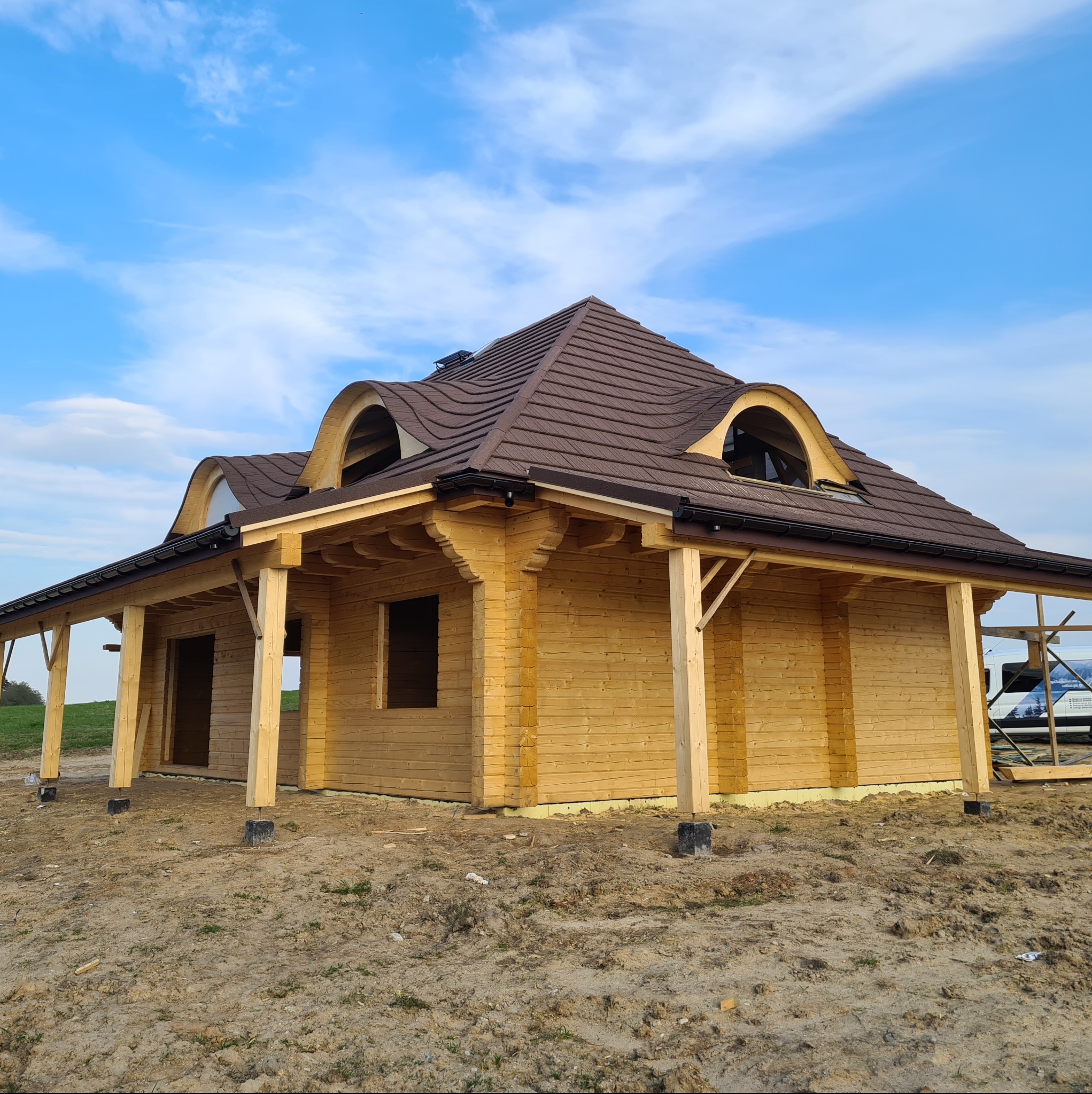 Niedokończony dom szkieletowy z brązowym dachem, widoczne drewniane elementy konstrukcyjne, okna dachowe w kształcie łuku i teren budowy.