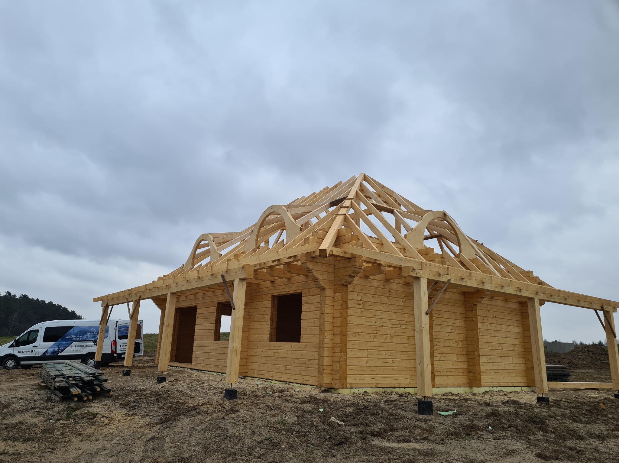 Surowy stan budowy domu z bali, widoczna konstrukcja drewniana dachu i ściany, na tle pochmurnego nieba, obok zaparkowany biały bus z logo firmy.