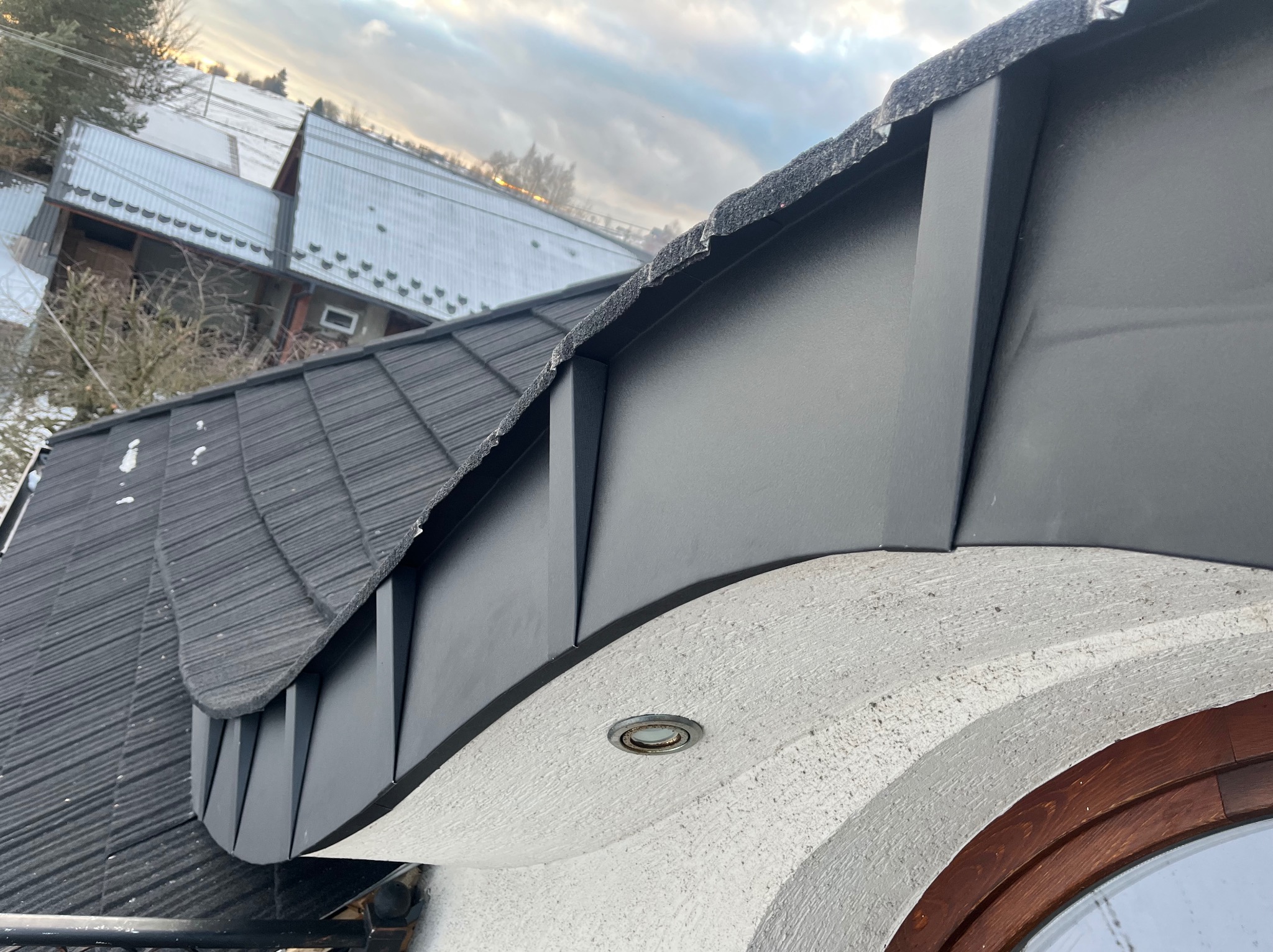Detale czarnego, metalowego wykończenia dachu z widocznym oświetleniem zewnętrznym wbudowanym w elewację budynku. W tle budynki pokryte śniegiem.