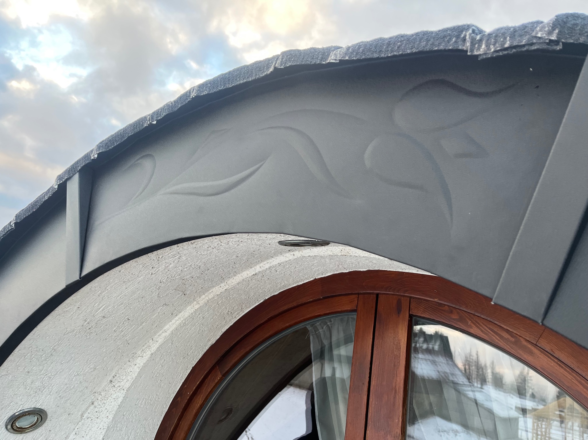 Szczegół dachu z ozdobnym grawerem nad okrągłym oknem z drewnianą ramą, widok na zaśnieżony krajobraz w tle.