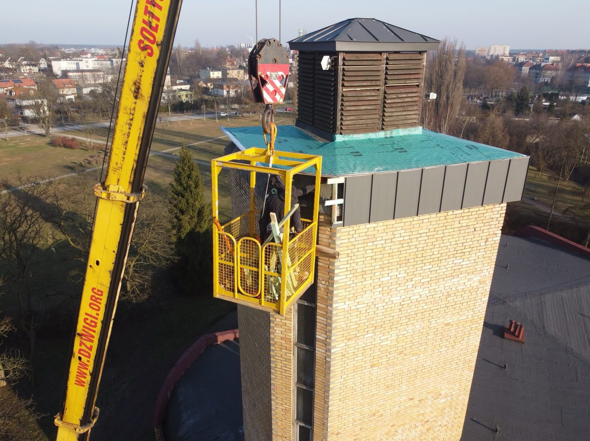 Pracownicy w żółtym koszu dźwigu pracują na wysokości przy wieży z cegły, na której znajduje się konstrukcja z drewna i blachy, widok na miasto w tle.