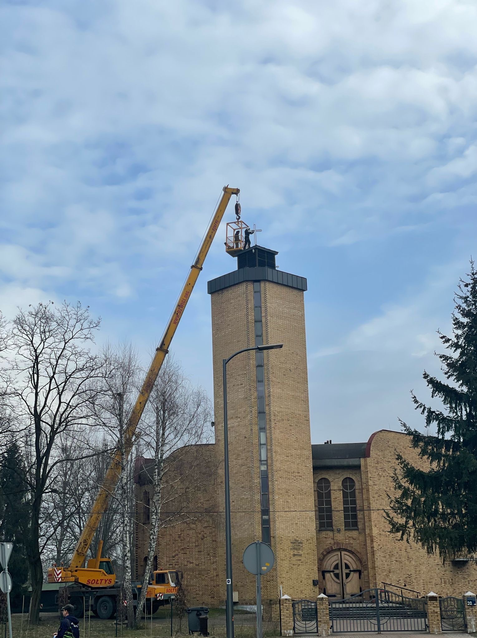 Prace na szczycie ceglanej wieży kościelnej wykonywane z podnośnika koszowego, widoczne narzędzia i robotnicy na wysokości.