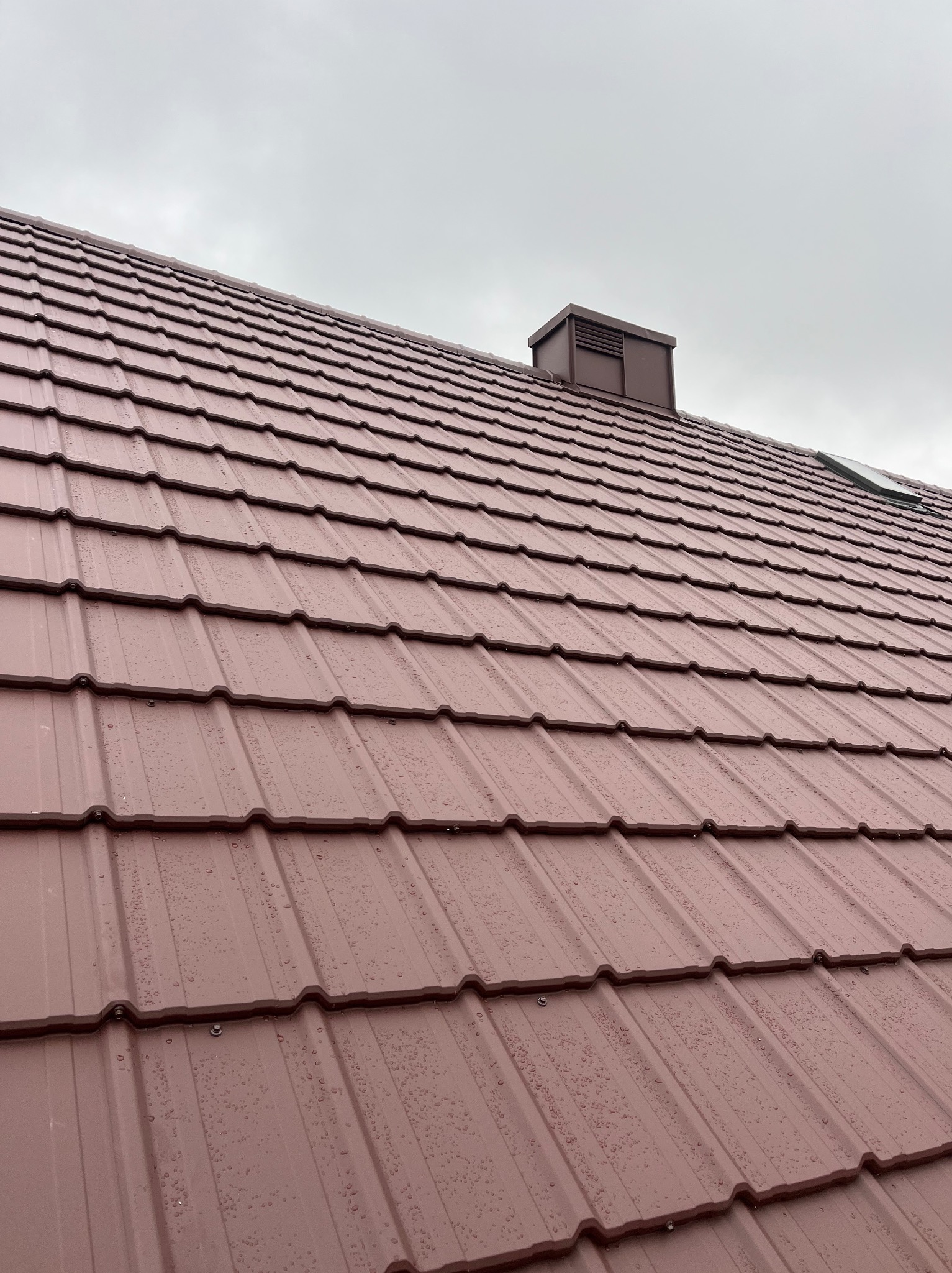Nowe, brązowe pokrycie dachowe z blachy trapezowej, widoczne krople deszczu i kominek wentylacyjny na tle pochmurnego nieba.