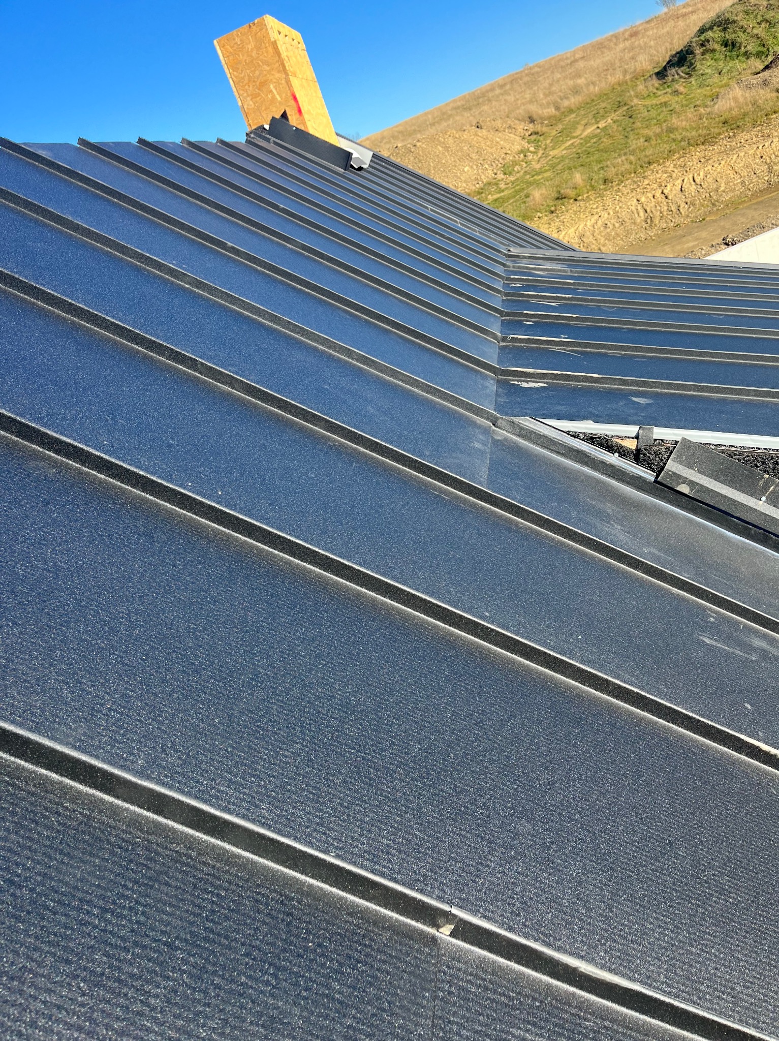 Nowe, czarne, metalowe pokrycie dachowe z widoczną konstrukcją komina w trakcie montażu na tle wzgórza i błękitnego nieba.